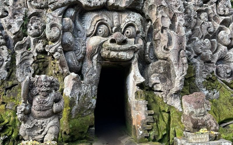 A stone temple entrance carved into a rock face, featuring a large, fierce face with wide eyes and an open mouth forming the doorway. Intricate carvings of figures and patterns surround the entrance. A moss-covered guardian statue stands to the left, and a small stone shrine with offerings sits to the right. The scene is weathered and lush, suggesting an ancient, sacred site in a tropical environment.
