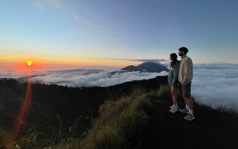 Two people standing on the grassy edge clifftop at the top of Mt Batur in Bali admiring the sunrise view and the clouds