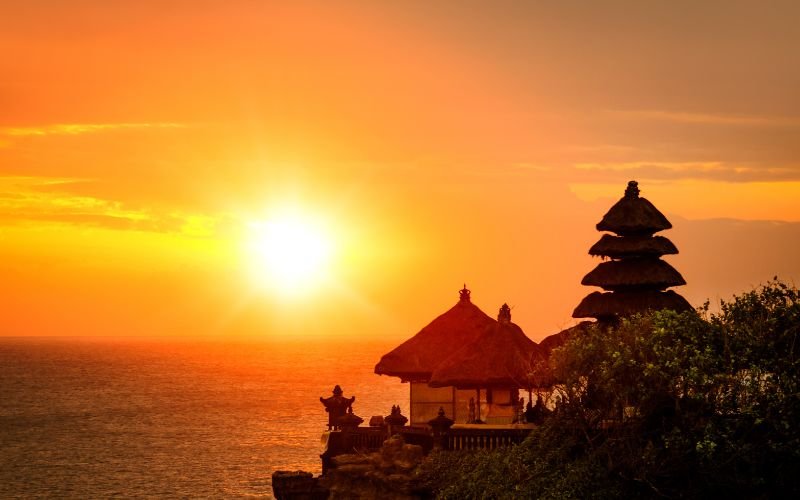 a beautiful bright orange and yellow sunset over the ocean and a clifftop temple with green trees