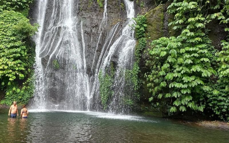 gushing waterfall leading into a big body of water with two people standing in it. The watefall is surrounded by lots of lush greenery