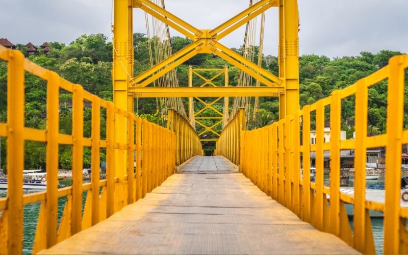 bright yellow metal bridge used by pedestrians and scooters with the ocean in the background surrounded by lush greenery