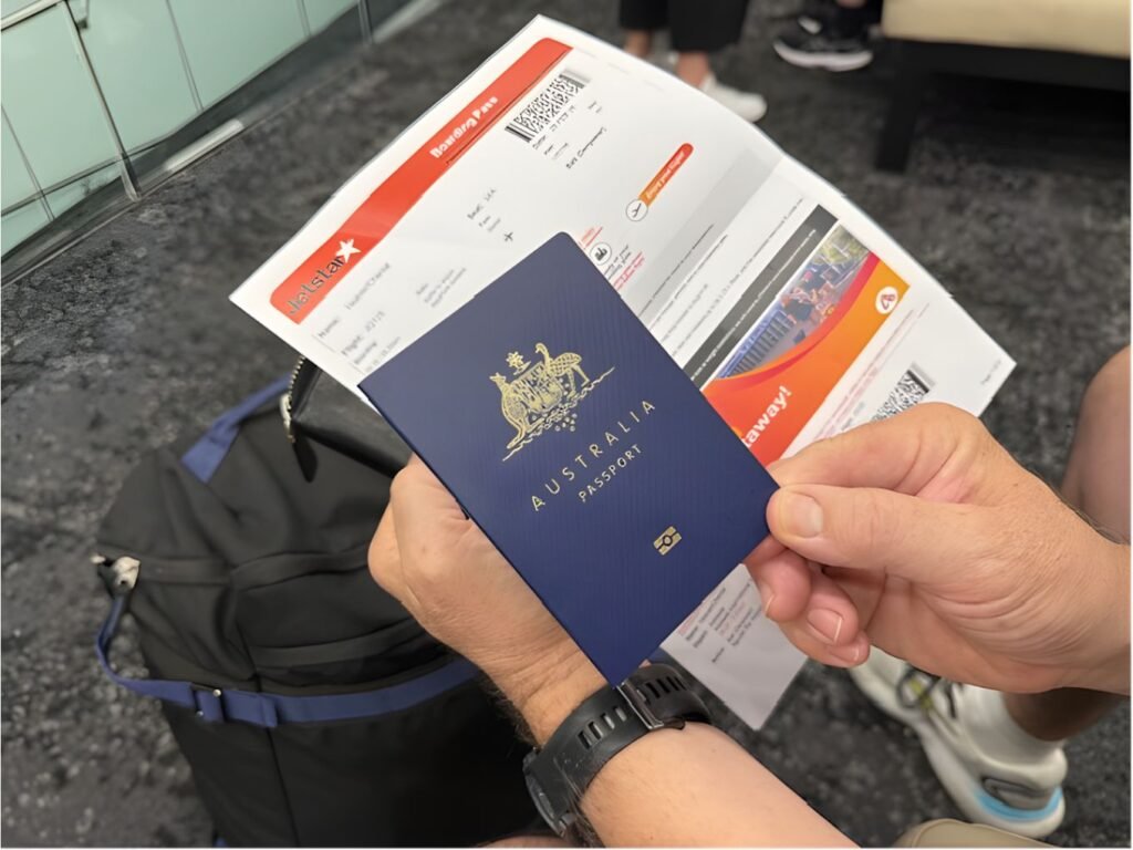 hand holding a blue Australian passport with a printed flight ticket and a black and blue backpack sitting on grey and black speckled carpet