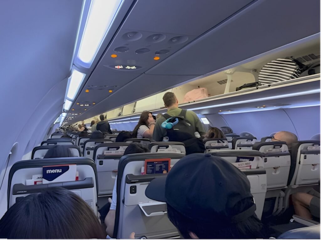 the inside of a dimly lit aeroplane cabin with some passengers seated and others standing in the aile. The overhead cabin bins are open displaying hand luggage
