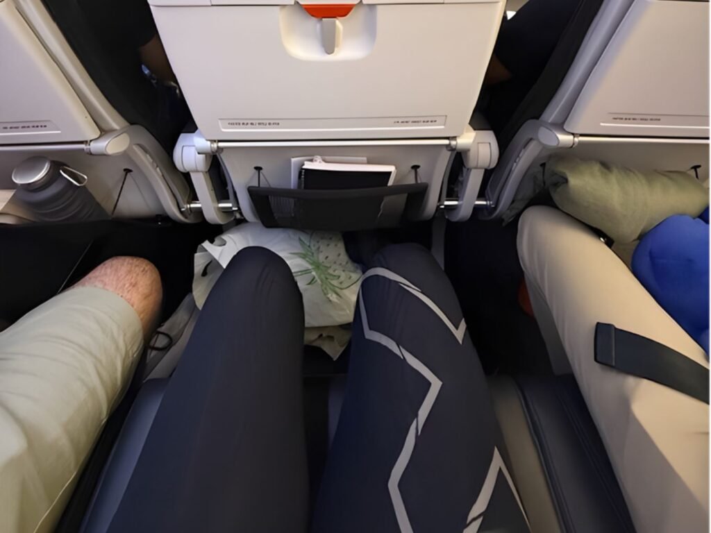 person seated in an aeroplane seat with the tray table stowed infront and a storage pouch underneath. It appears the person does not have a lot of legroom