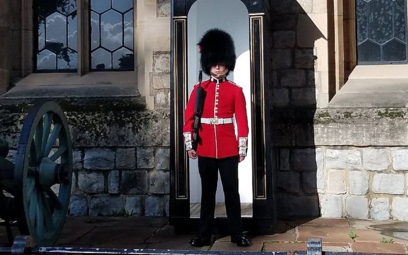 British guard wearing black pants and shoes with a red and white jacket holding a gun on his side and a big black fur hat standing infront of a stone building with decorative windows