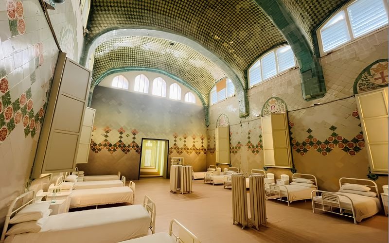 antique hospital beds set up as an exhibition in a room with a tiled dome arch ceiling and lino flooring with window shutters behind the beds