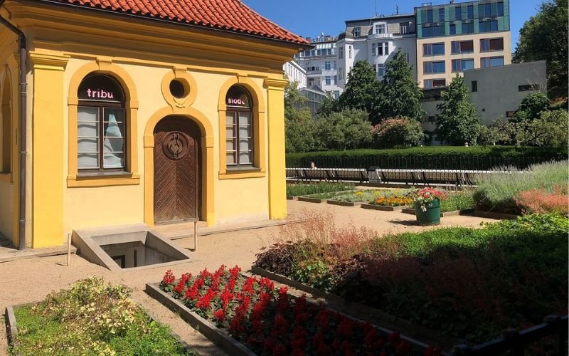 neatly manicured garden with red flowers and green hedging and grass with a quaint yellow building with a red tiled roof behind it