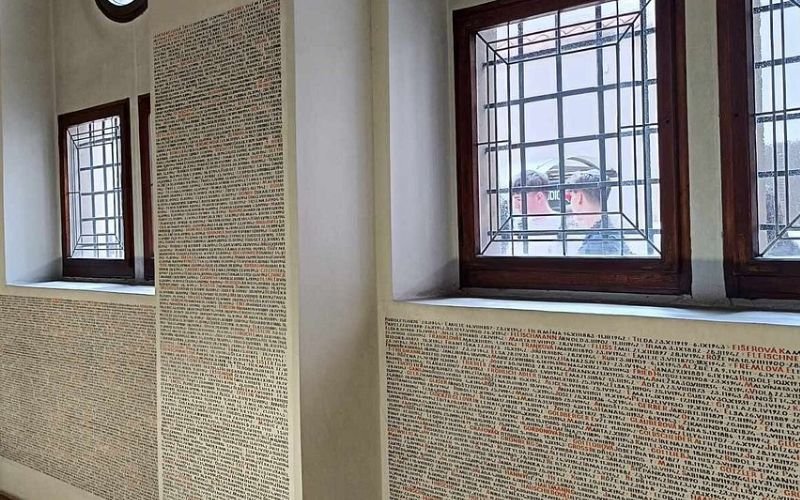 interior of a synagogue featuring thousands of names of people in tiny black writing along the walls with a glass window in the background