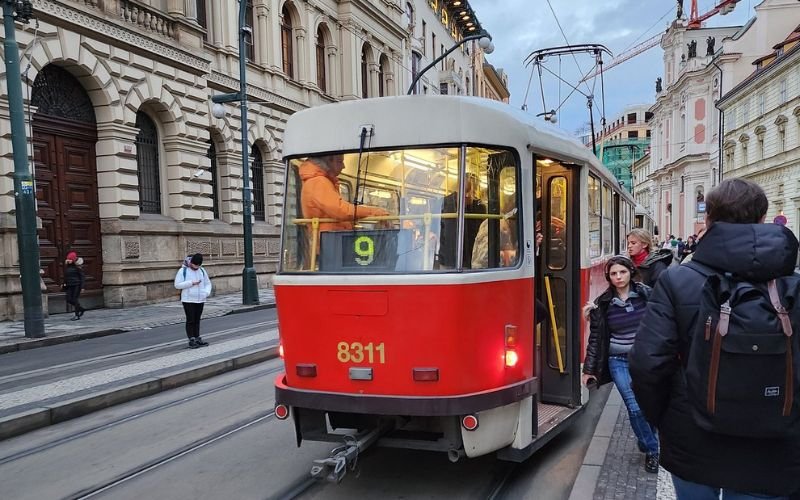 red and white tram stopping at tram stop as people board surrounded by a historic building with arched windows